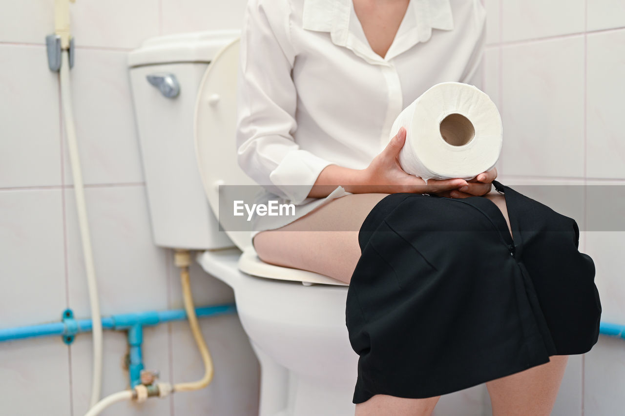 Midsection of woman sitting in bathroom with toilet paper at home