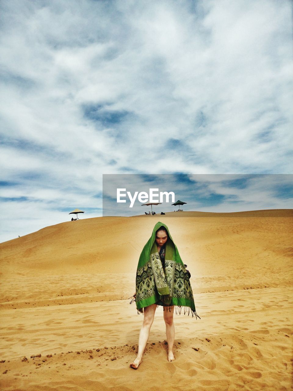 Full length of woman wrapped in shawl standing on sand at beach against cloudy sky