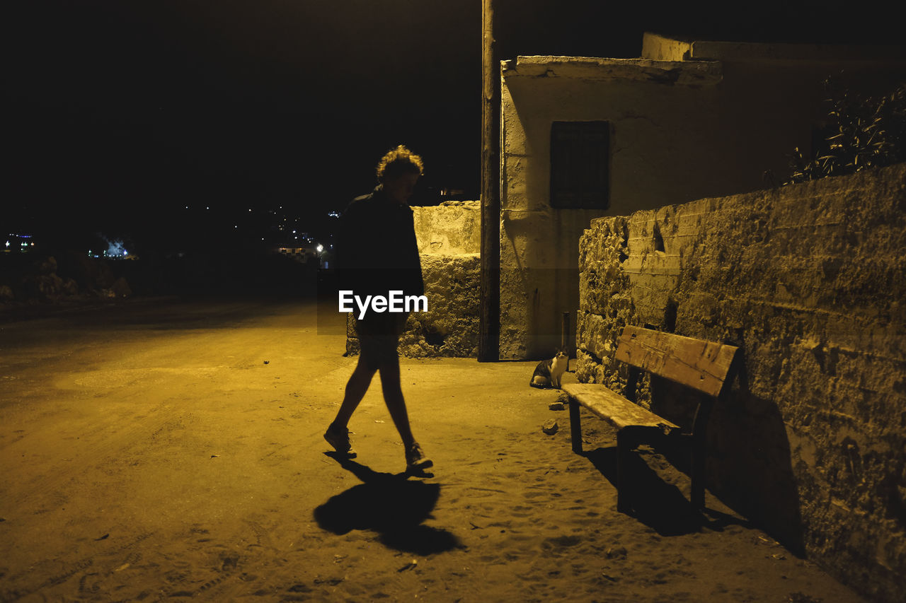 FULL LENGTH REAR VIEW OF MAN STANDING ON STREET AGAINST BUILDING AT NIGHT