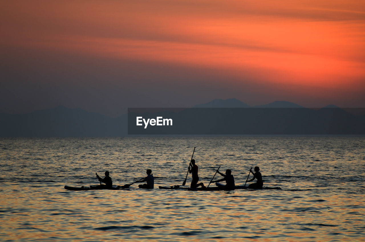 Silhouette people paddleboarding in sea against orange sky