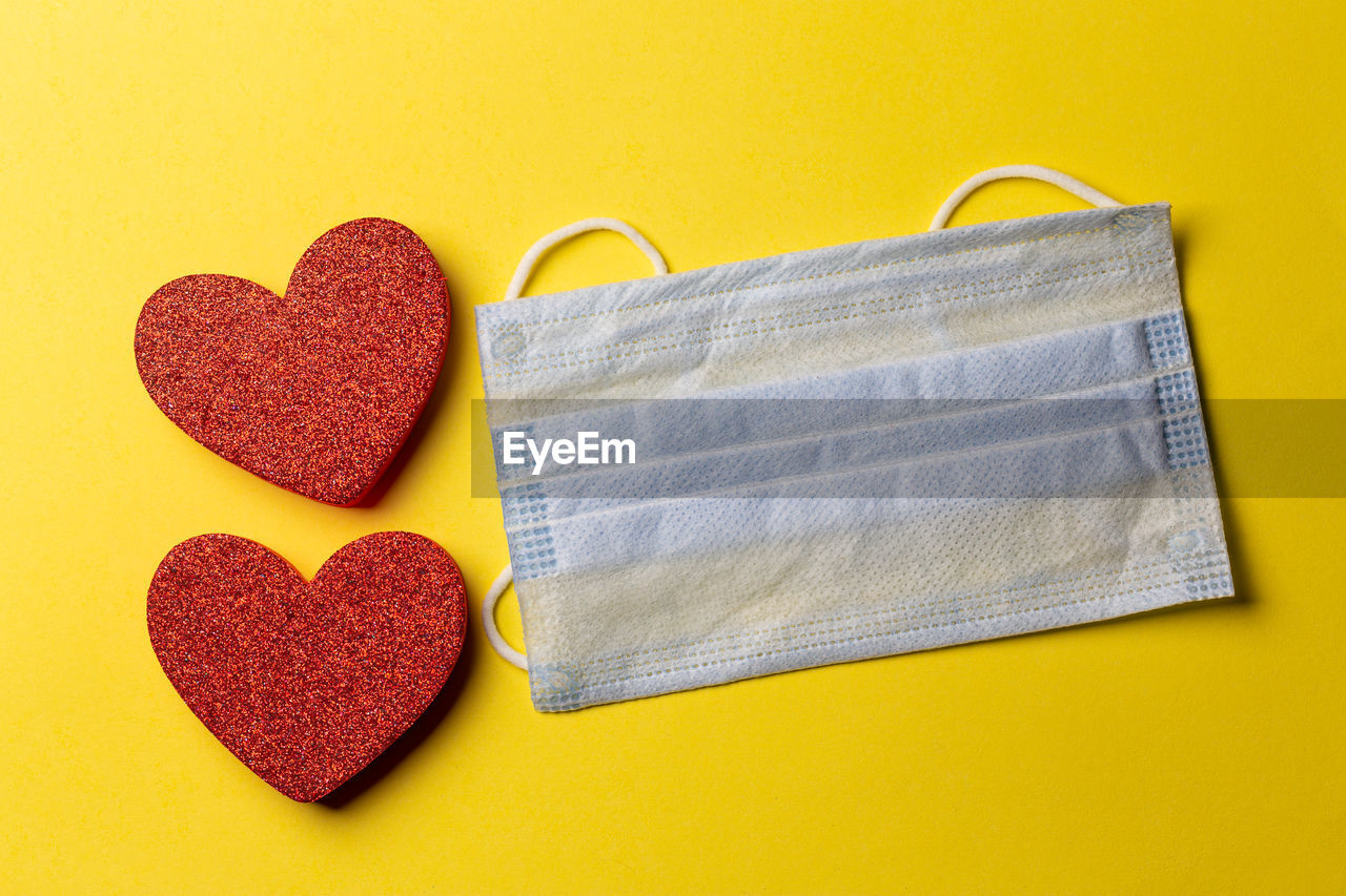 Top view of two red heart-shaped and a medical face mask on a yellow paper background