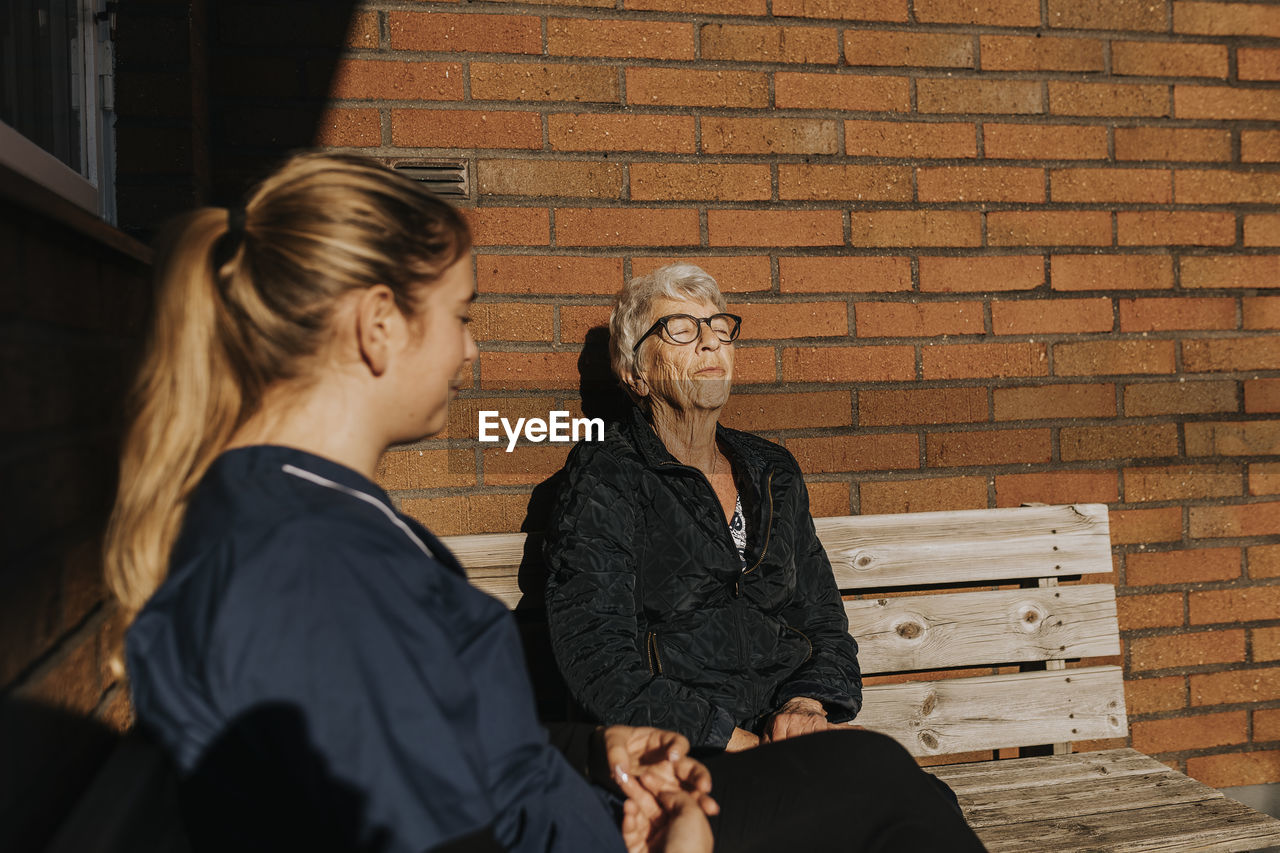 Home caretaker and senior woman sitting on bench and sunbathing