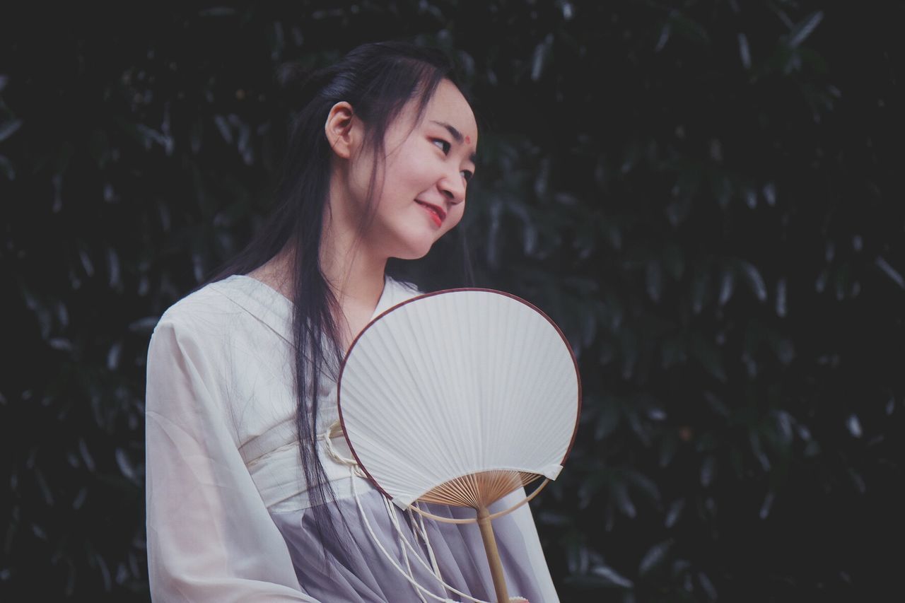 Smiling woman with hand fan standing against plants