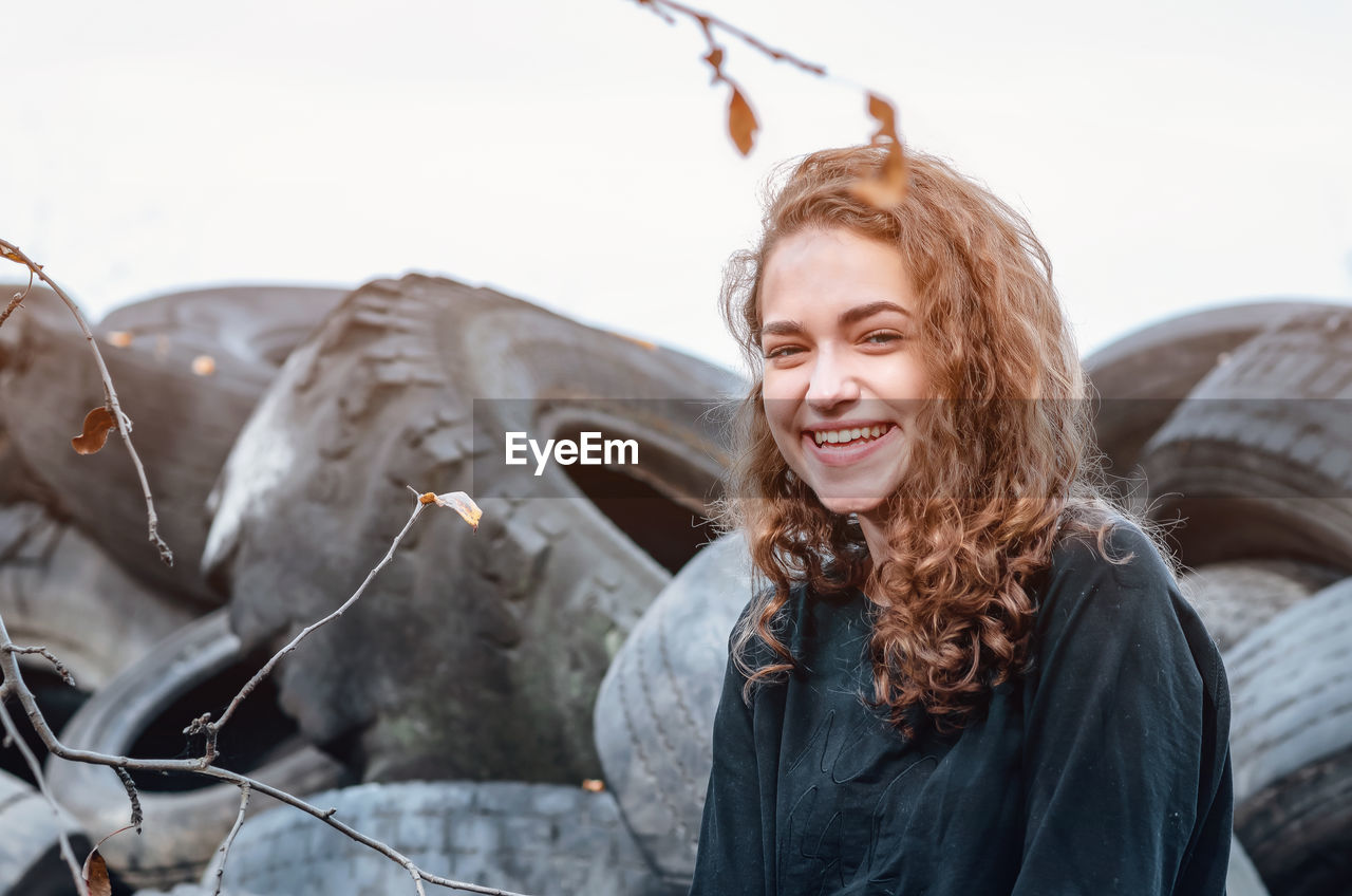 Cheerful curly-haired girl standing against a landfill of used car tires