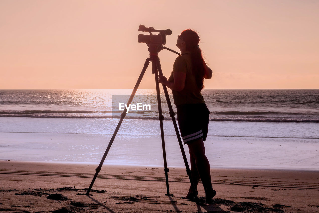 Side view of woman standing with movie camera on tripod at beach