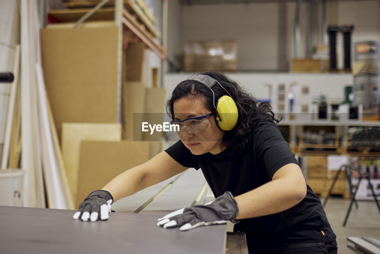 Serious female carpenter wearing protective eyewear and ear protectors while working in industry