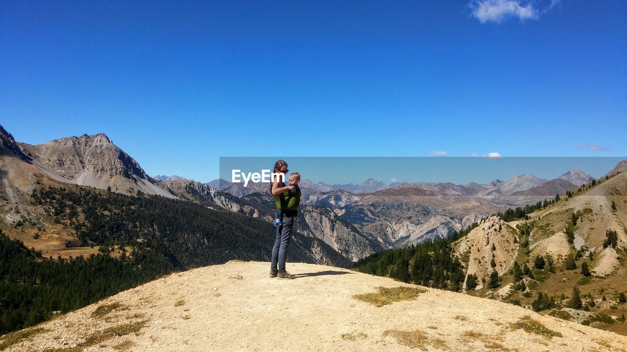 Mother carrying son while standing on top of mountain against blue sky during sunny day