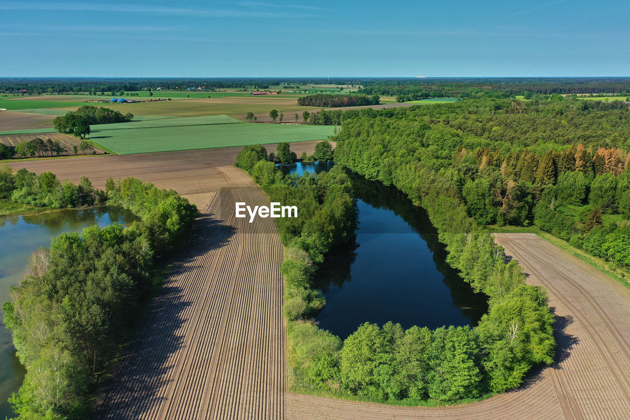 Aerial view of a small dark blue pond with high trees on the bank between fields