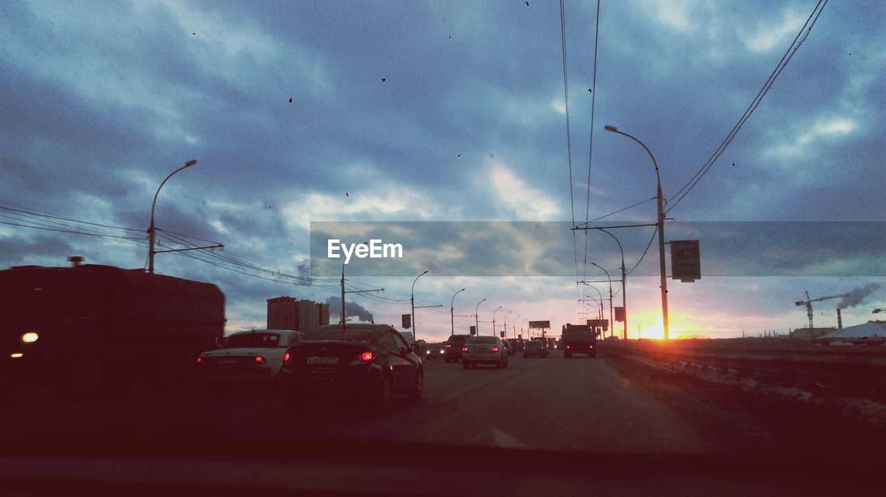 Cars on street against cloudy sky seen through windshield during sunset