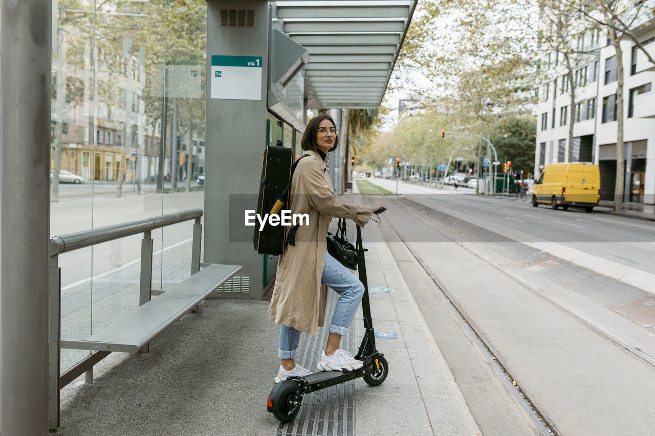 Woman with push scooter and instrument case standing at tramway station in city