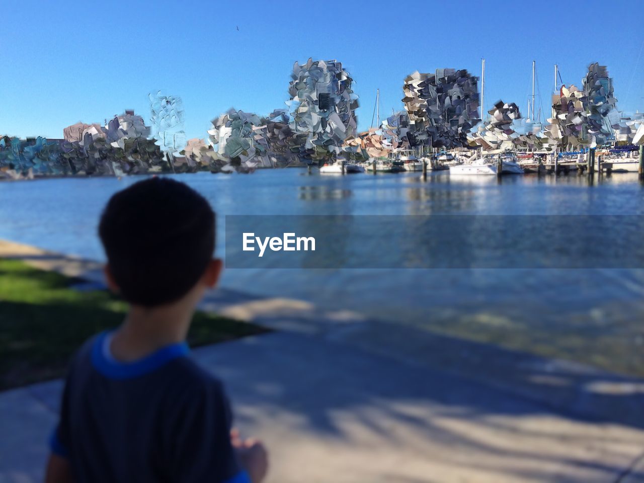 Rear view of boy looking at paper structure by river against clear blue sky