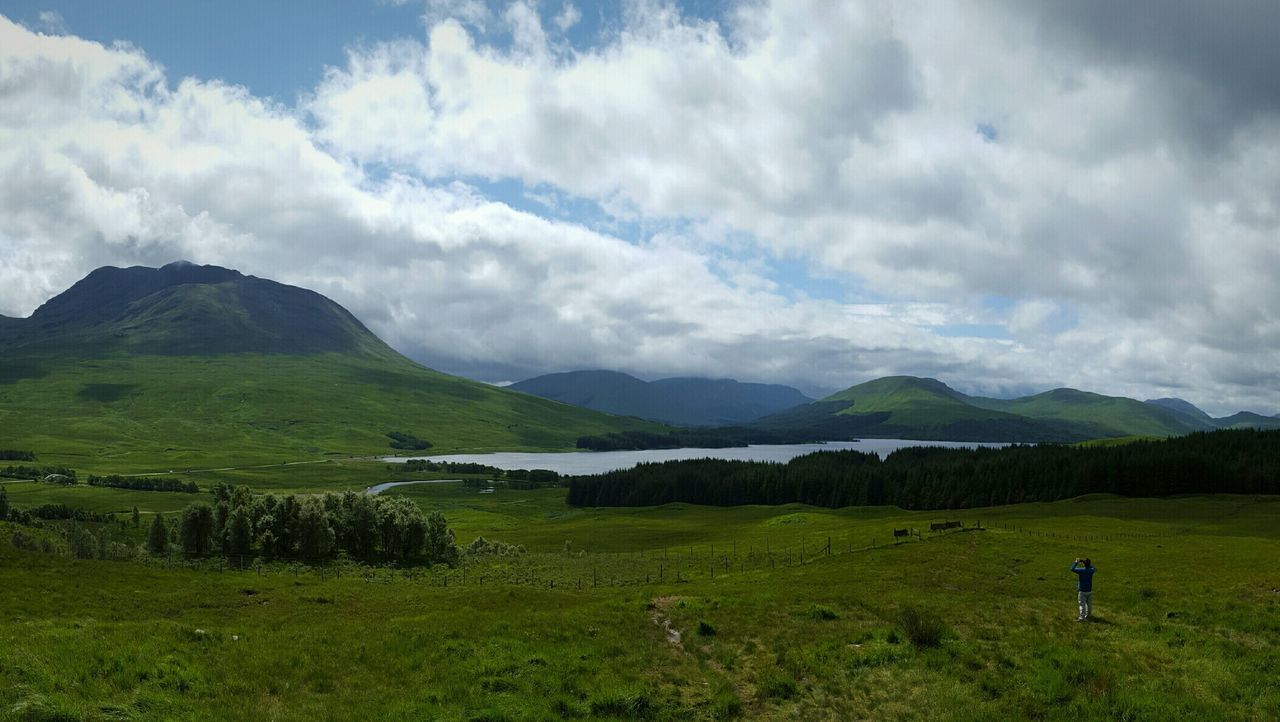 Man standing on grassy field by lake and mountains against sky