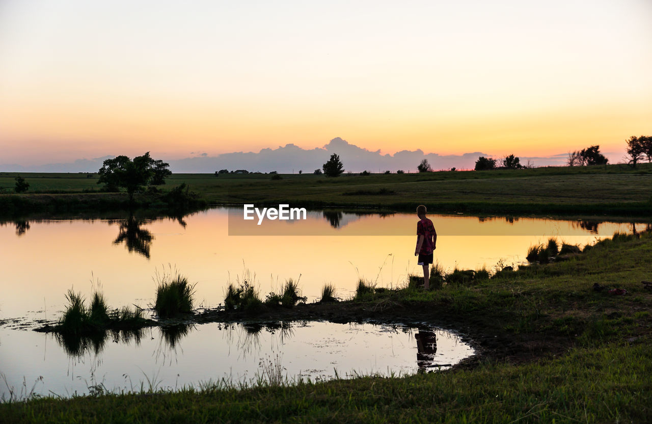 Boy by pond against clear sky during sunset