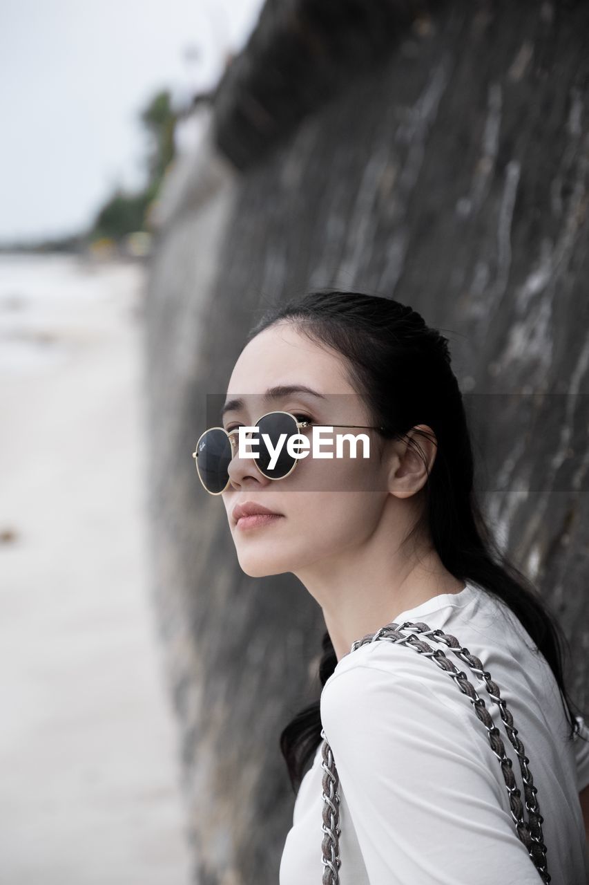 Young woman wearing sunglasses while standing by stone wall at beach