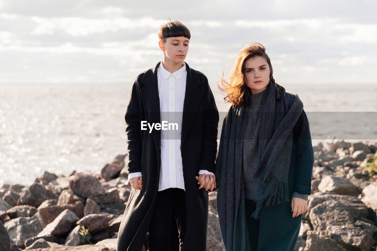 Lesbian women holding hands while standing on rock against sea and sky