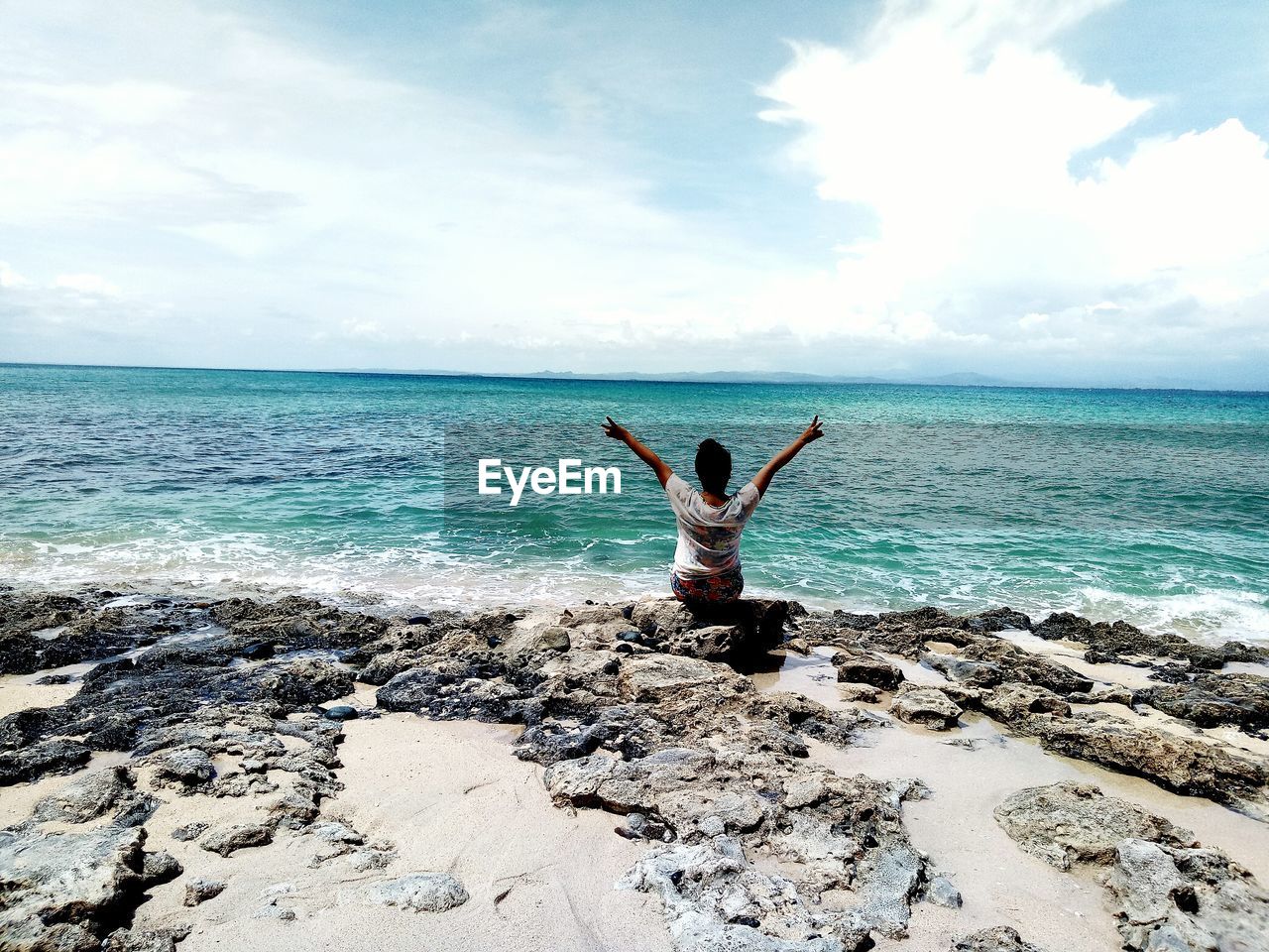 Rear view of woman gesturing peace sign while sitting on shore against sky