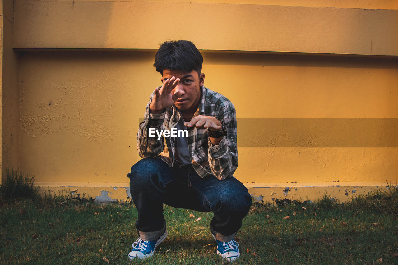 Full length portrait of young man gesturing while crouching against wall