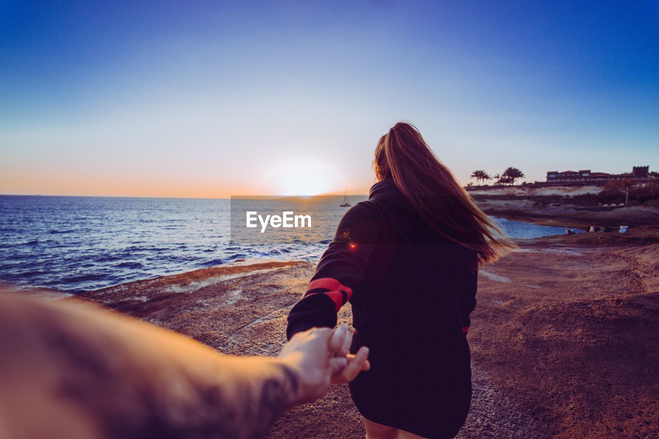 Cropped image of man holding woman hand at beach during sunset