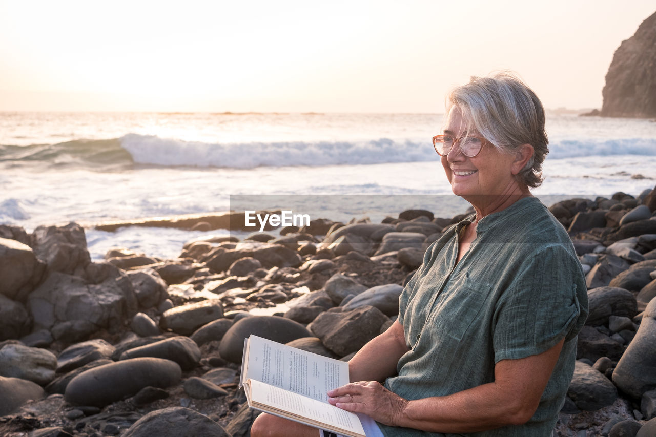 FULL LENGTH OF MAN SITTING ON ROCK AT BEACH AGAINST SKY