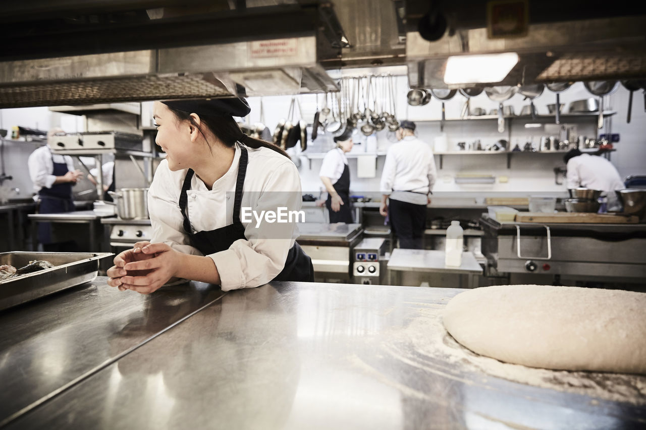 Female chef leaning on counter with colleagues in background at commercial kitchen
