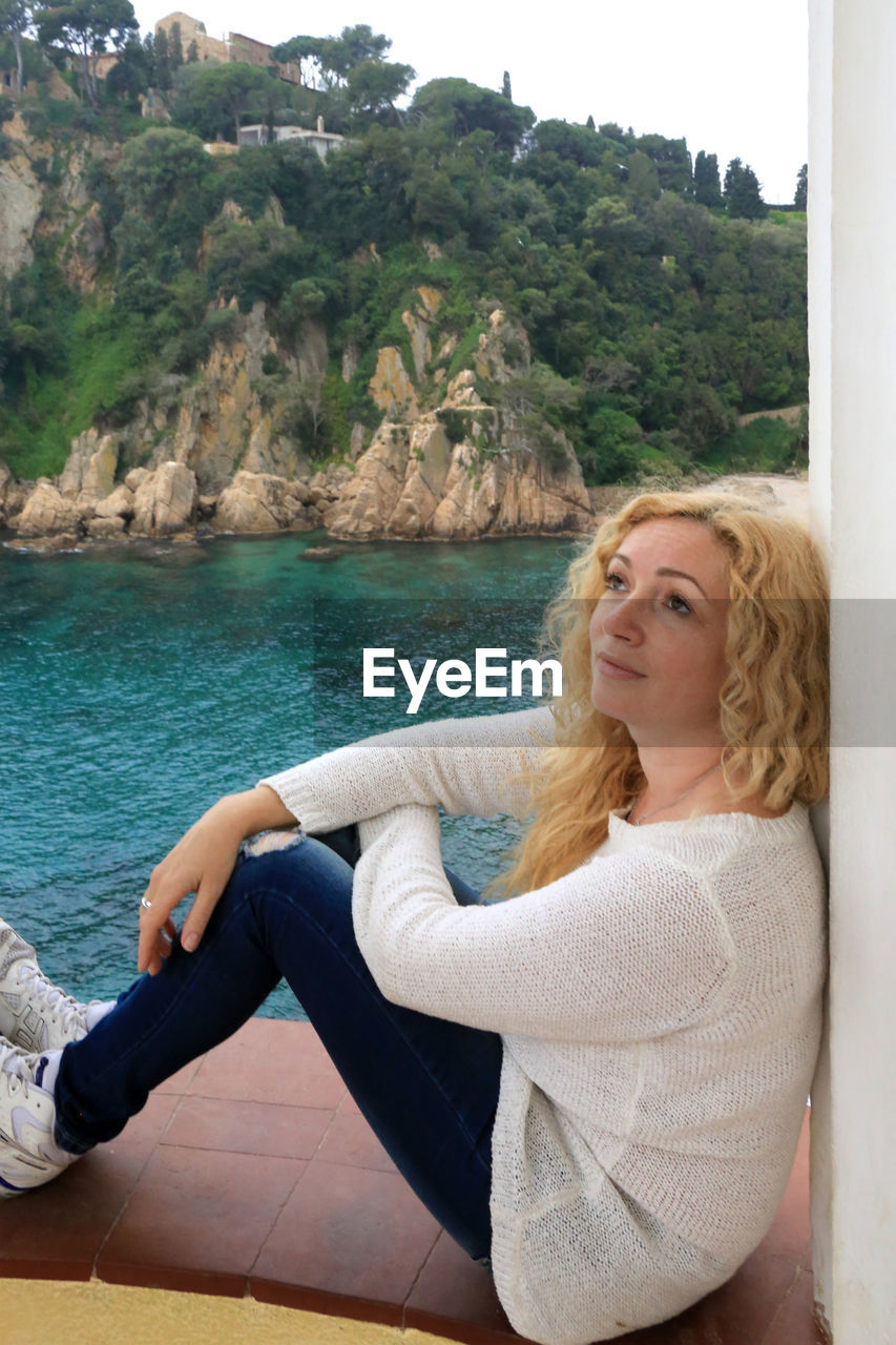 PORTRAIT OF BEAUTIFUL YOUNG WOMAN SITTING AGAINST SEA