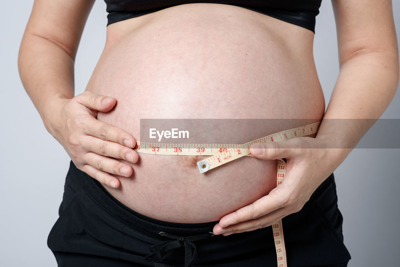 Midsection of pregnant woman measuring stomach with measuring tape over gray background