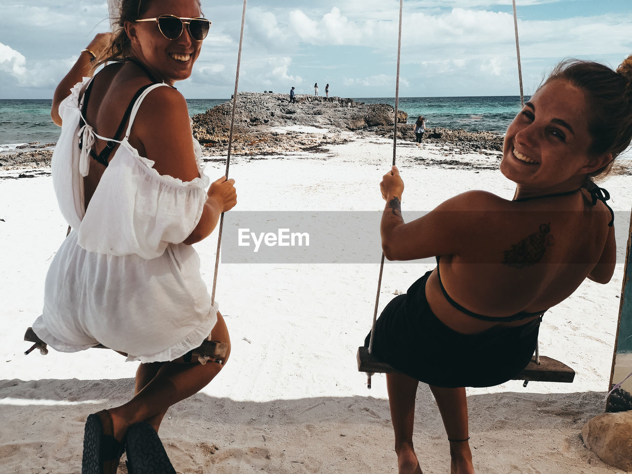 Cheerful of friends sitting on swing at beach against sky