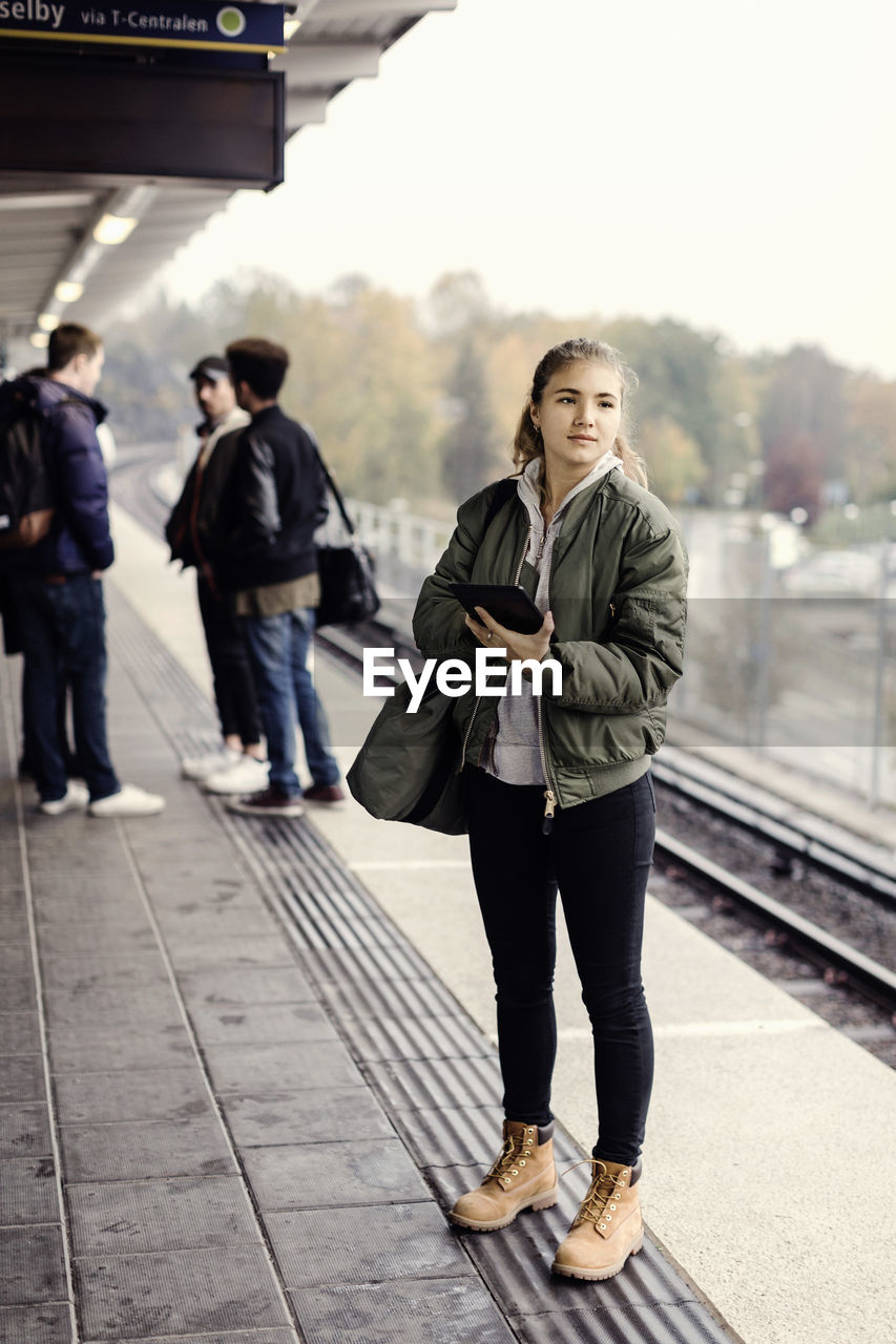 Full length of female university student holding digital tablet at subway station with friends in background