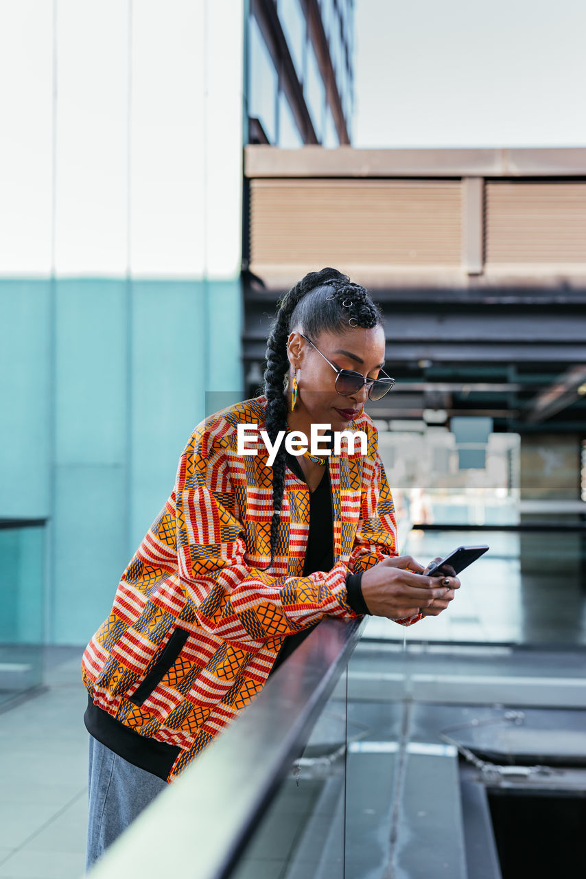 Side view of young ethnic female in stylish colorful clothes leaning on glass fence and browsing social media on smartphone on terrace outside modern building