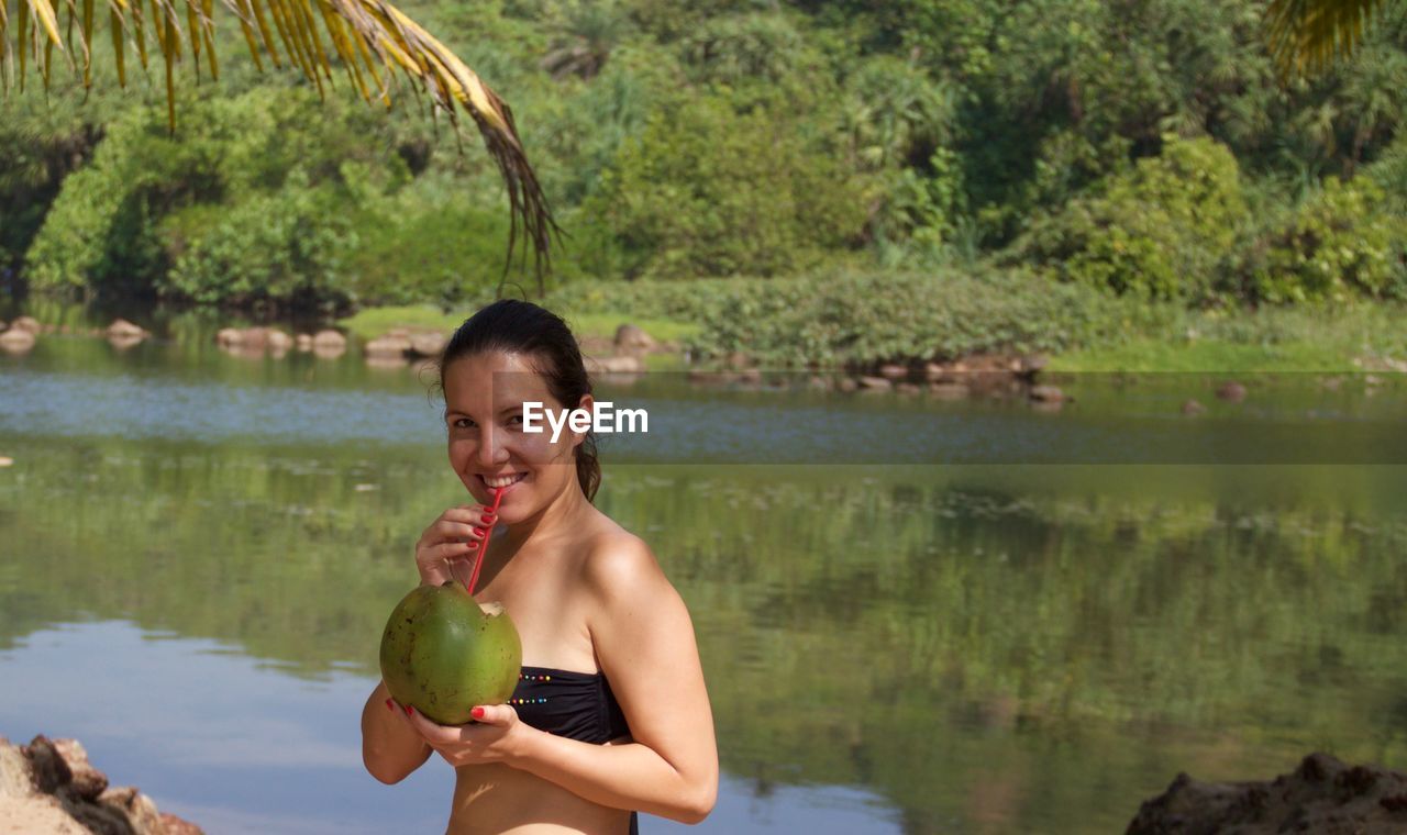 Portrait of smiling young woman drinking coconut water by lake at arambol