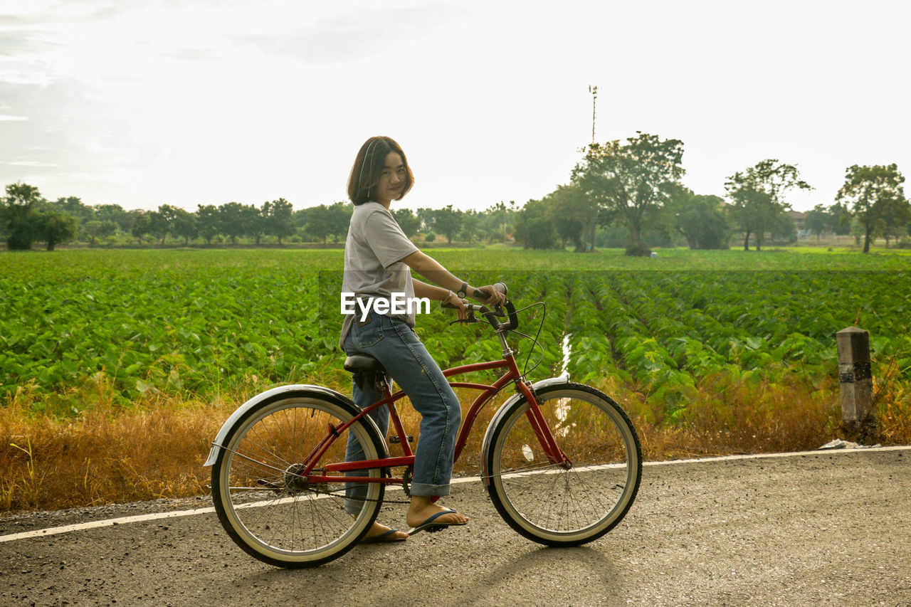 Portrait of young woman with bicycle on road by farm against sky