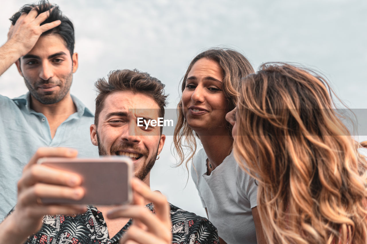 Close-up of cheerful young man showing smart phone to friends against sky