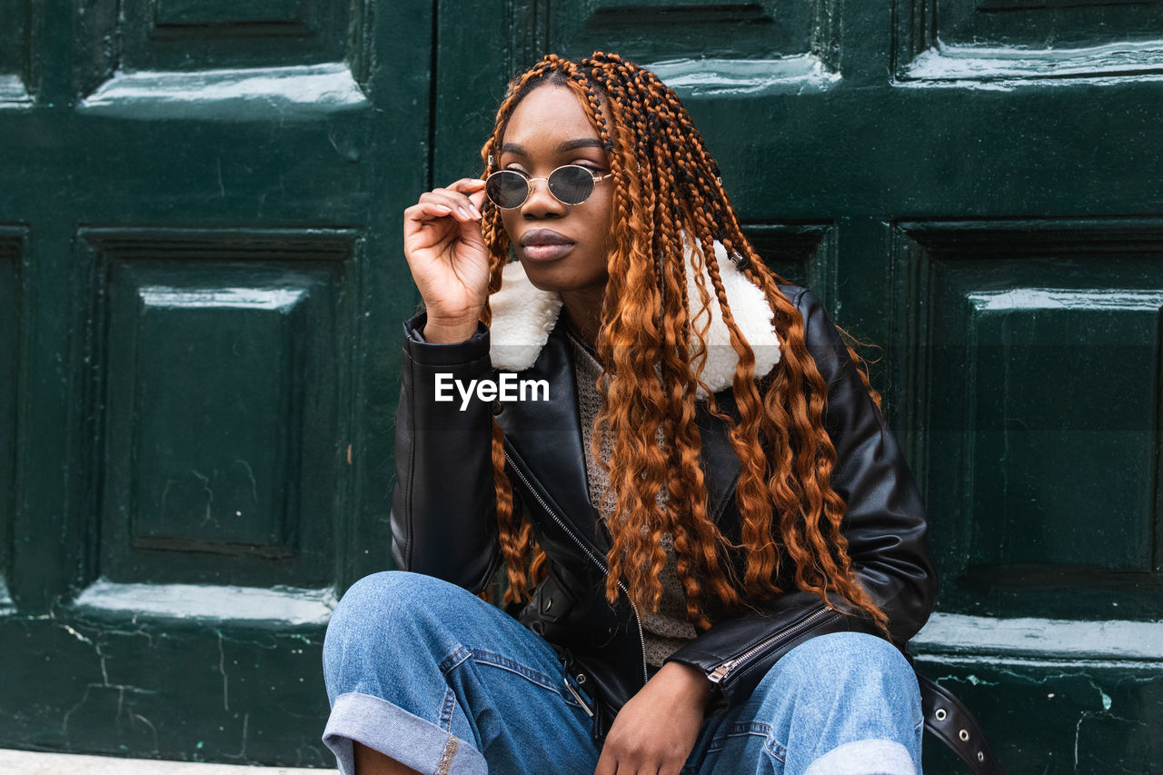 Confident african american female in trendy outfit and sunglasses with long braided hair looking away in city street