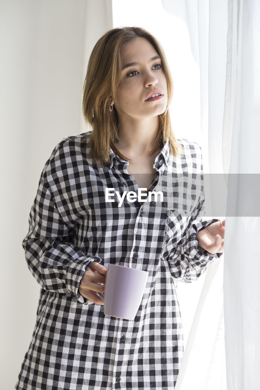 Young woman holding coffee cup by curtains at home