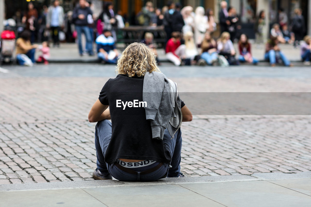 Rear view of young man sitting on footpath on crowded street