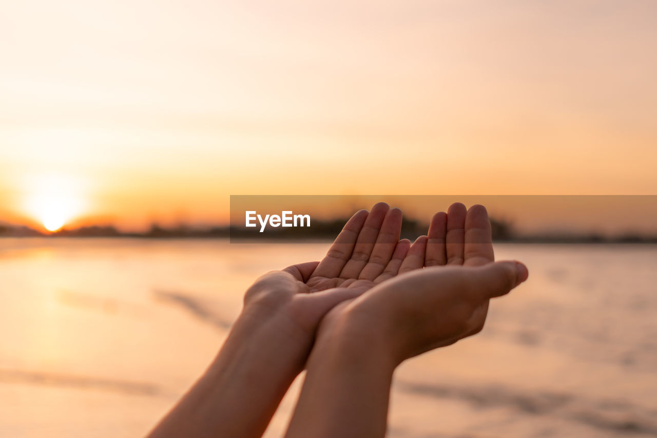 Midsection of woman hand by sea against sky during sunset