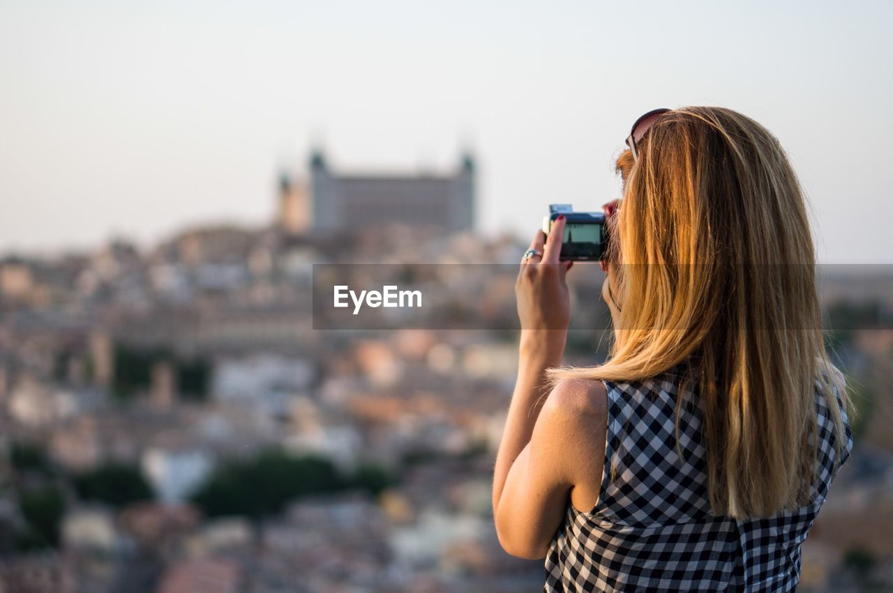 Rear view of girl photographing buildings using camera on sunny day