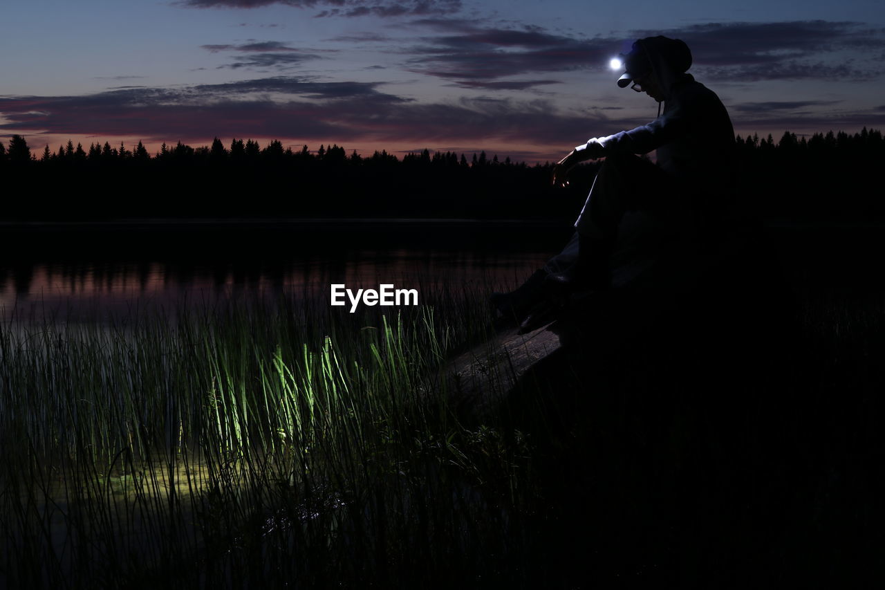 Silhouette man sitting with illuminated flashlight by lake against sky during sunset