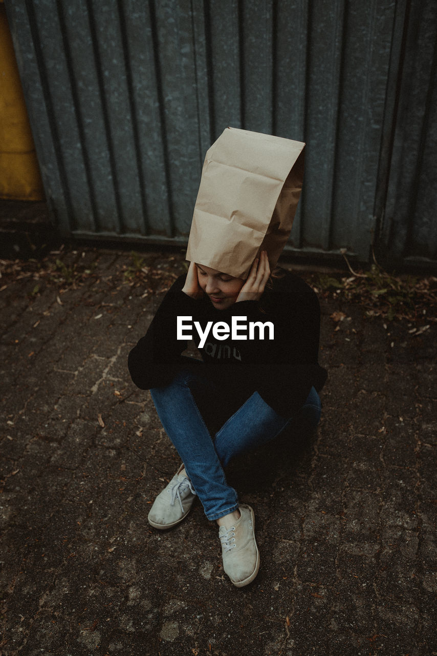 High angle view of woman wearing paper bag sitting against wall