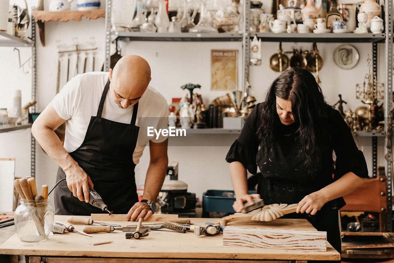 Concentrated mature artisan man and woman with professional tools creating wooden details while working together in art workshop