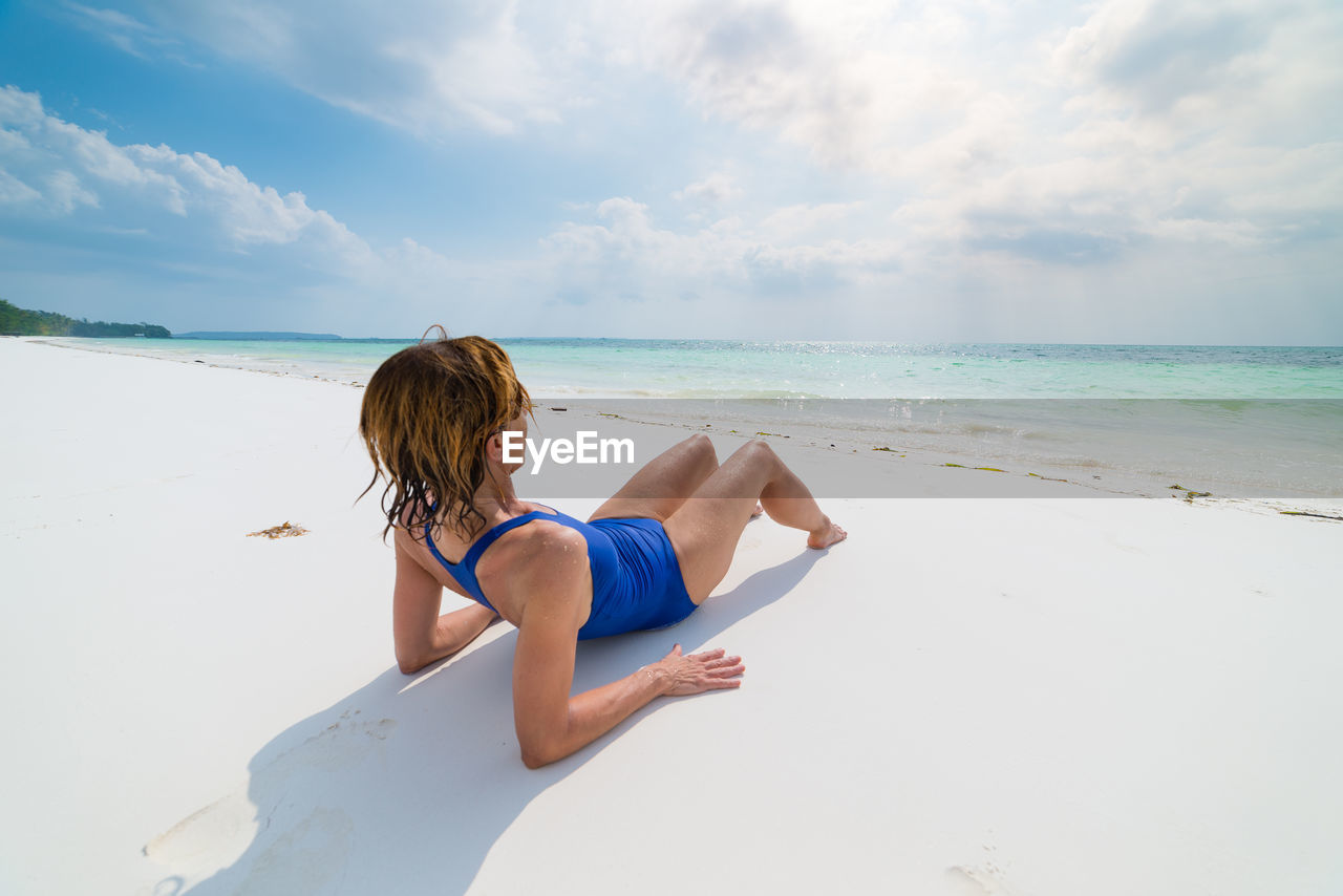 Mature woman wearing swimwear lying on beach against cloudy sky during sunny day