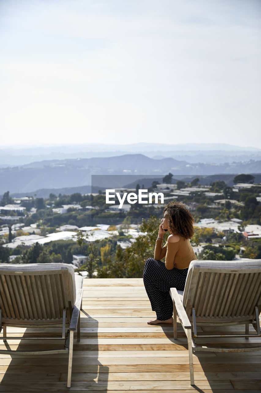 Side view of woman talking on mobile phone while sitting on deckchair against sky