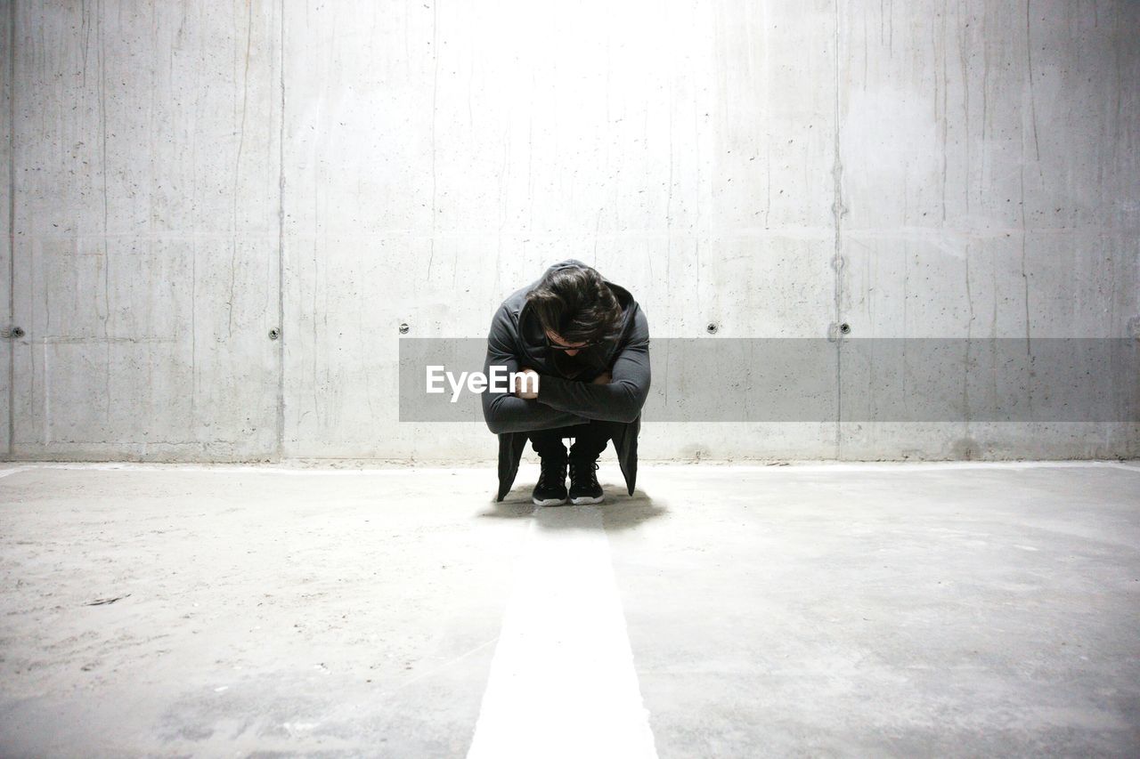 Full length of sad man crouching in illuminated underground parking lot