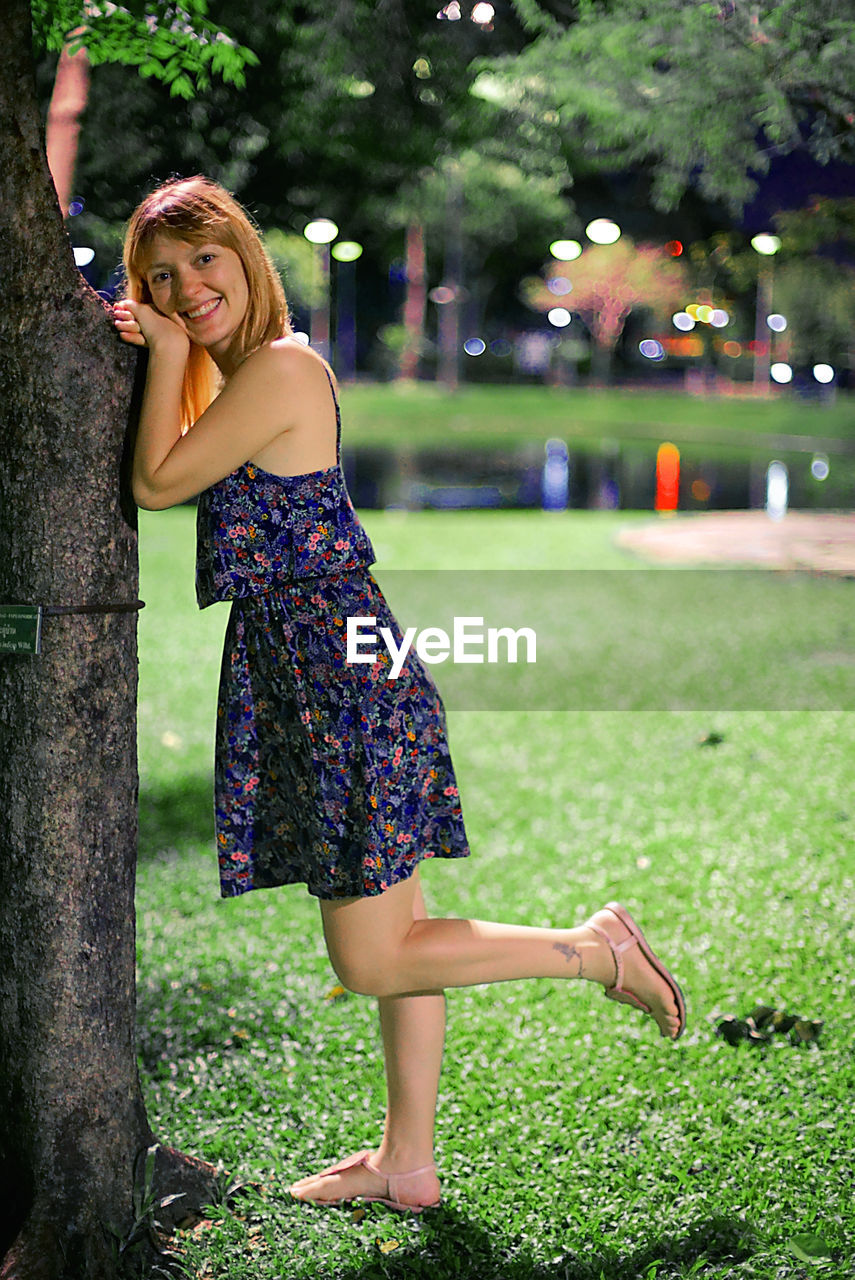 FULL LENGTH PORTRAIT OF SMILING YOUNG WOMAN AGAINST TREES