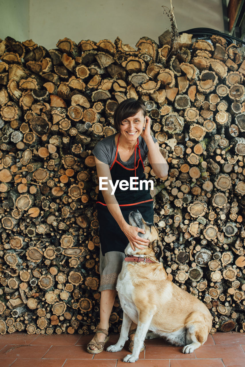 Smiling middle age woman in apron with dog at pile of sawn logs