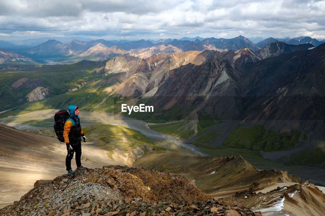 REAR VIEW OF MAN LOOKING AT MOUNTAIN RANGE AGAINST SKY