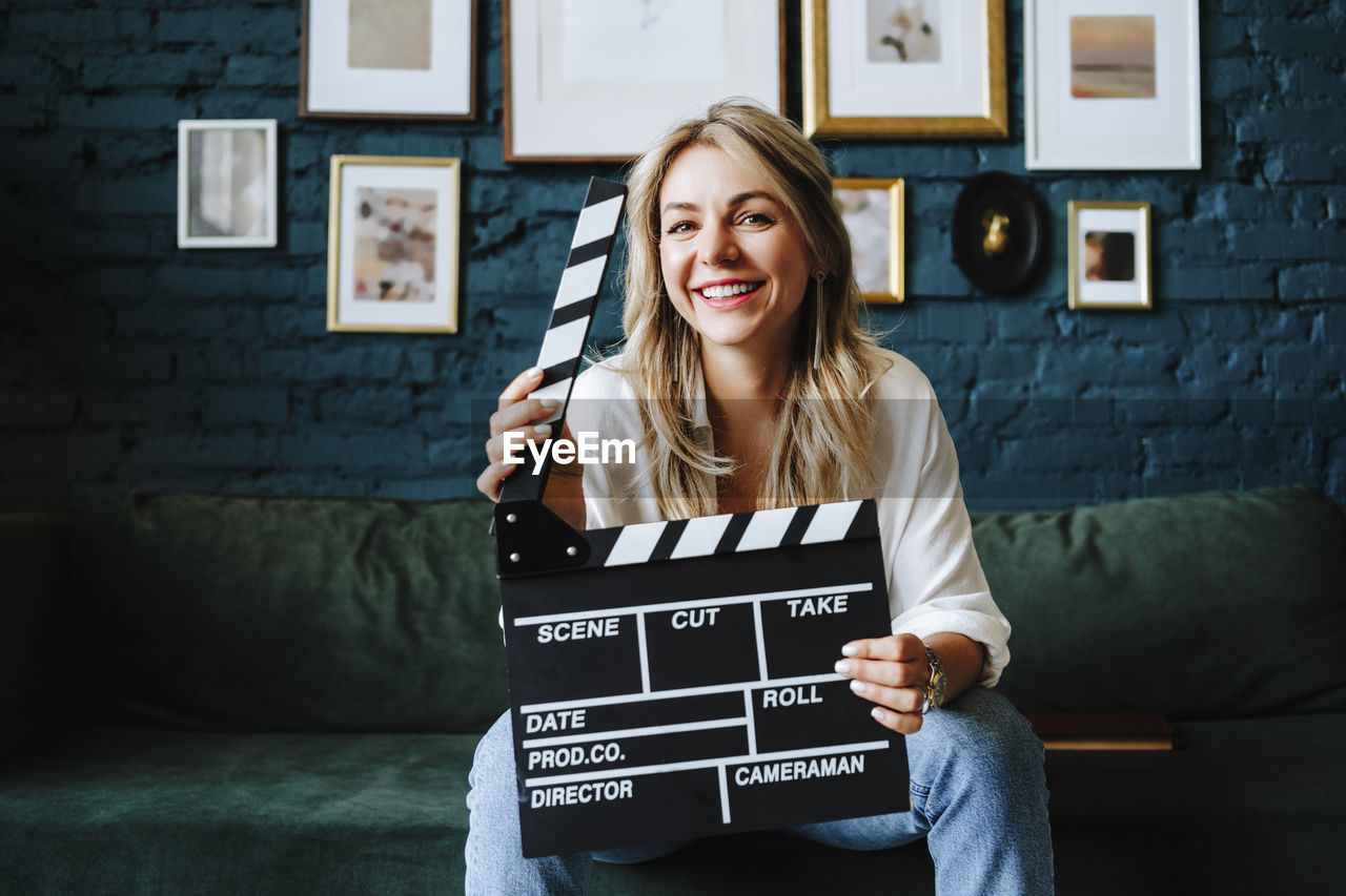 Happy actress holding clapboard sitting on sofa at film set