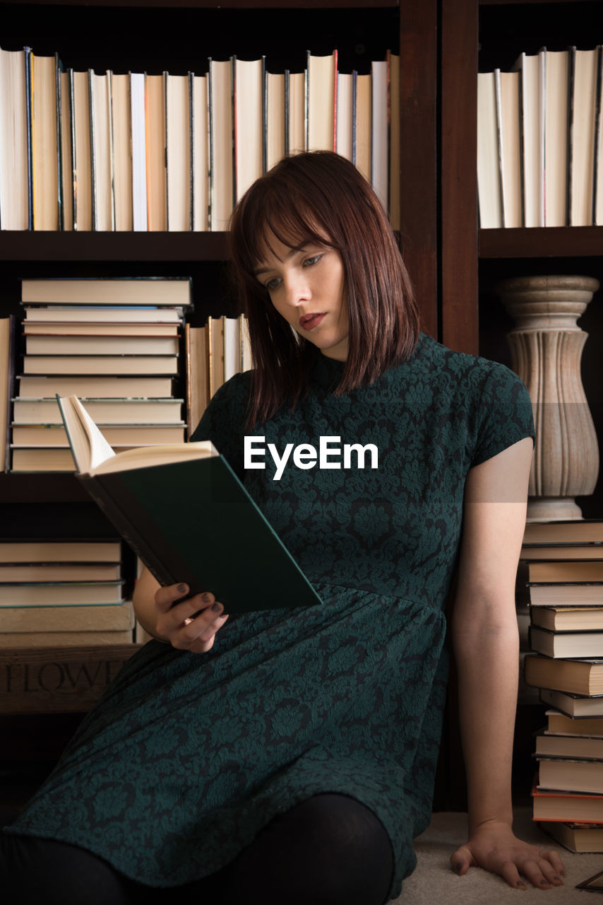 Young woman reading book while sitting against bookshelf in library