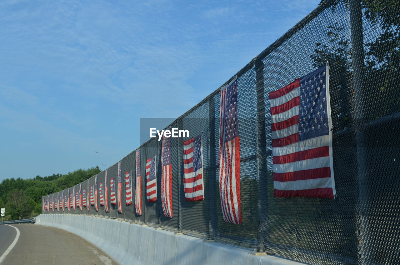 Row of american flags on fence of highway overpass on sunny side of road, with road and white line