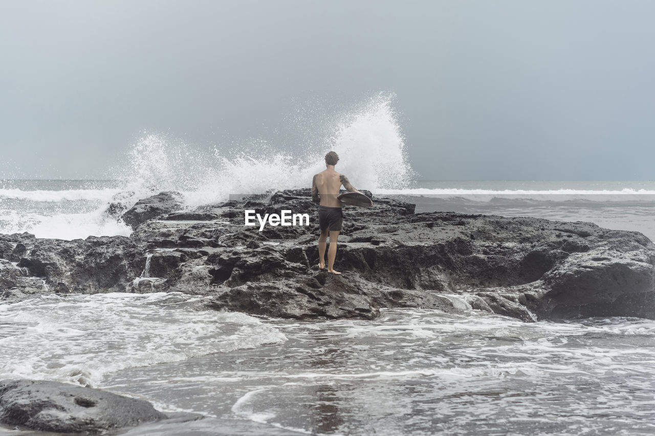 Rear view of man carrying surfboard while standing on rocks at beach against sky